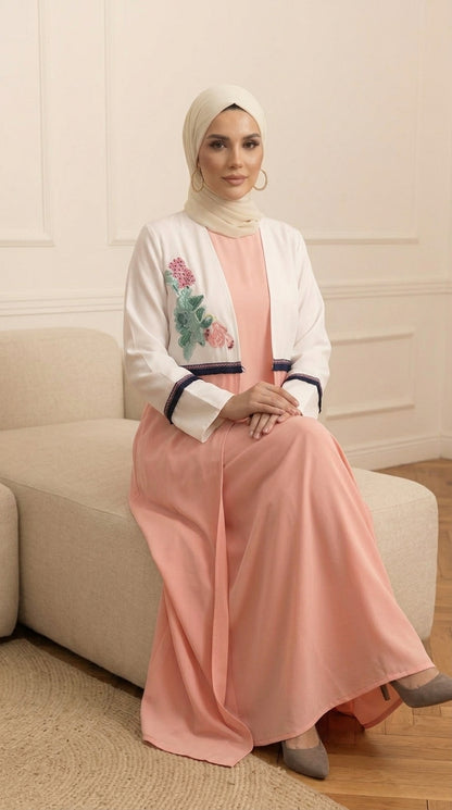 A woman wearing a peach-colored maxi dress with a white embroidered jacket and a cream hijab, sitting on a beige sofa in a neutral room.