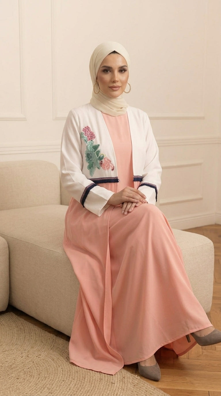 A woman wearing a peach-colored maxi dress with a white embroidered jacket and a cream hijab, sitting on a beige sofa in a neutral room.