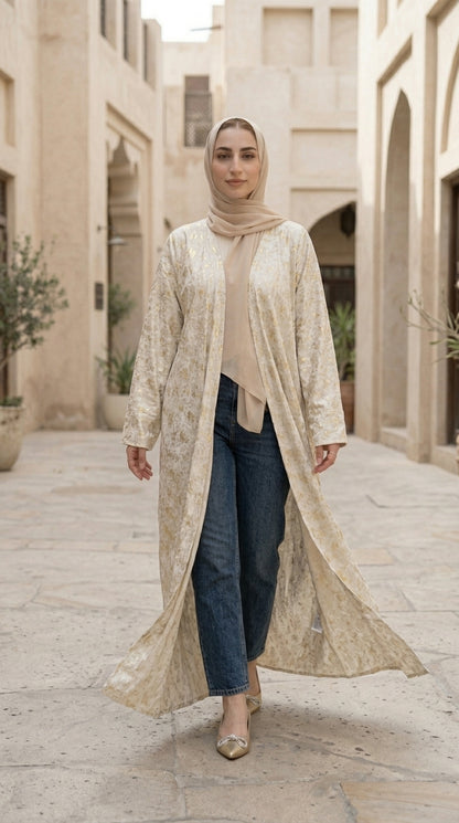 Woman wearing a long beige embroidered jacket in an outdoor setting with architectural elements.