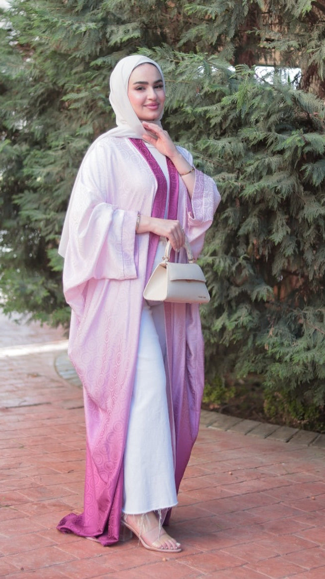 A woman wearing a light pink and purple ombré abaya with a matching hijab stands on a brick pathway in front of a green tree, holding a small beige handbag. A 'RAFE' brand watermark is visible in the bottom right corner.