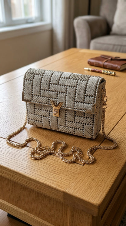A structured woven handbag in light beige and gray with a gold-tone Y-shaped turn-lock clasp and a gold chain strap, resting on a wooden table in a cozy indoor setting.