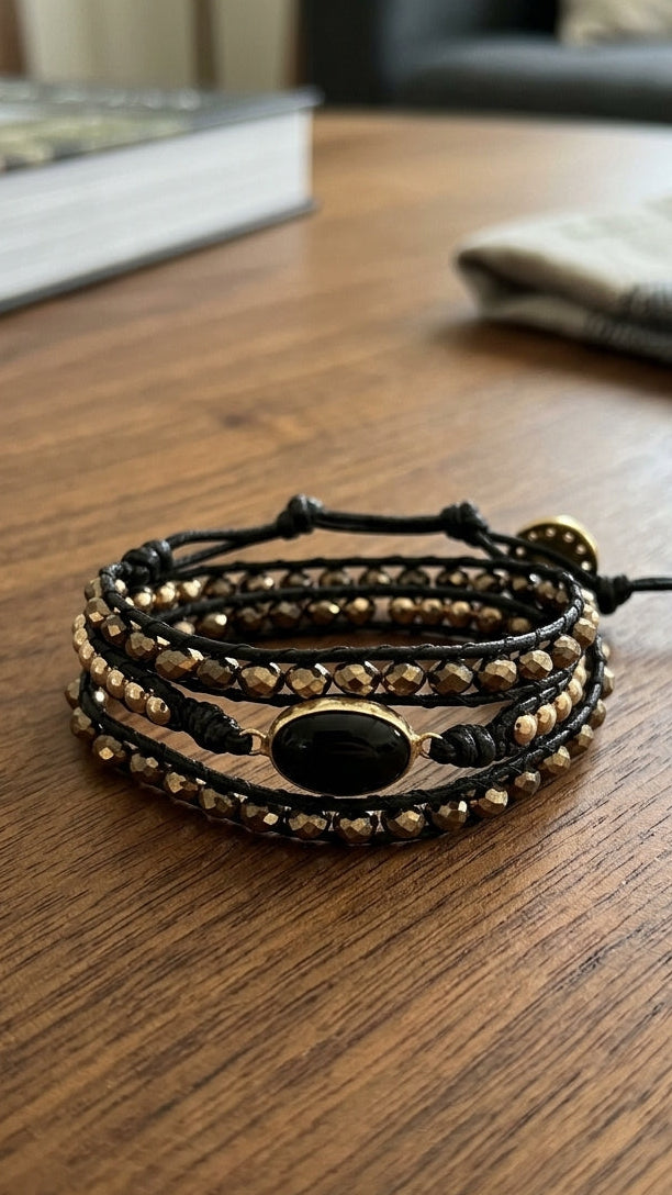 A multi-strand black leather wrap bracelet with gold-toned beads and a central black oval stone, resting on a wooden table.
