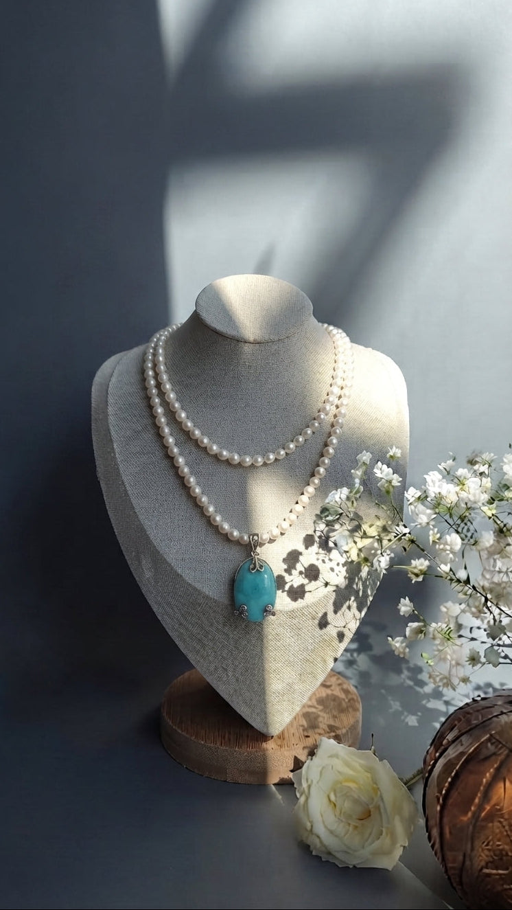 A double-strand pearl necklace with a turquoise pendant displayed on a textured cream jewelry bust. The bust is set on a wooden base with decorative white flowers and a brown textured sphere on a dark gray surface, lit by soft natural light with shadows.