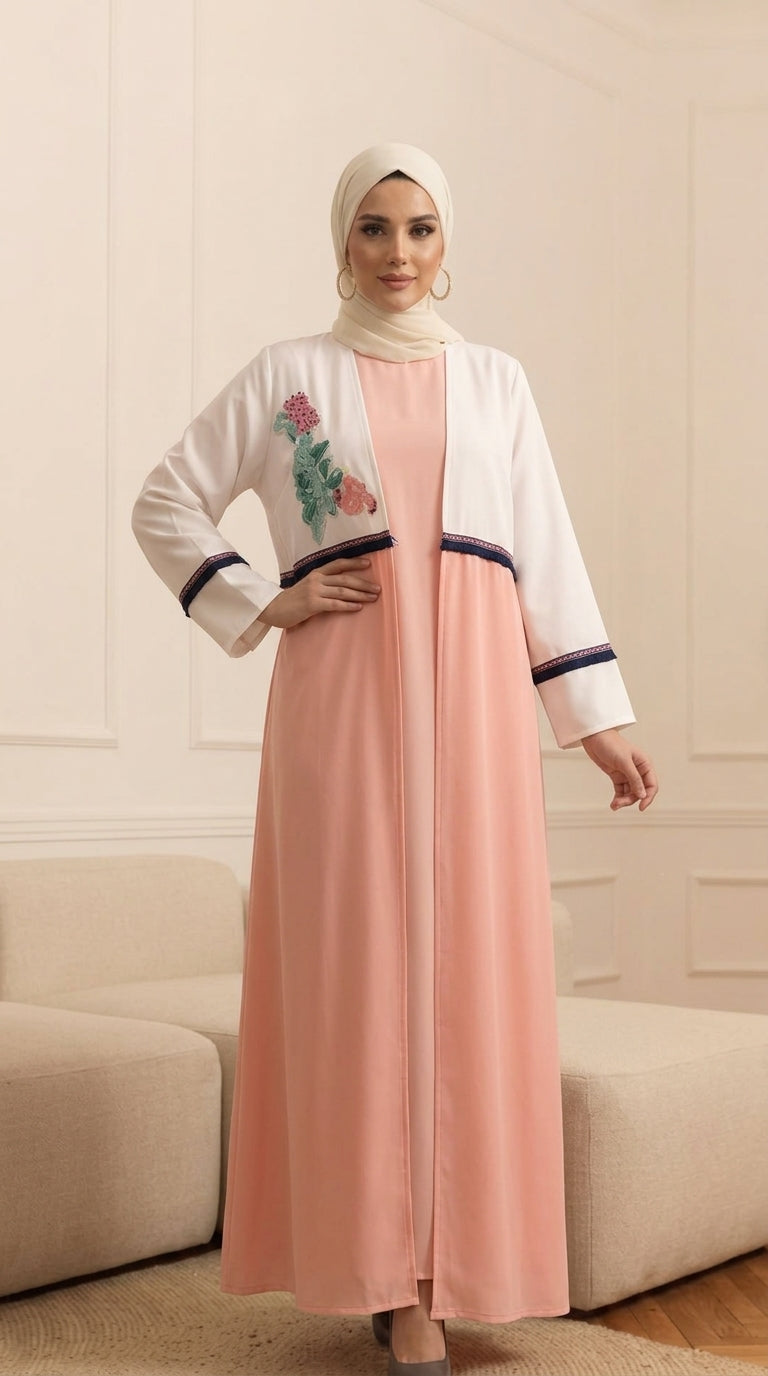 A woman wearing a modest outfit consisting of a peach-pink abaya dress with a white embroidered jacket and a cream hijab, standing in a neutral-toned indoor setting.