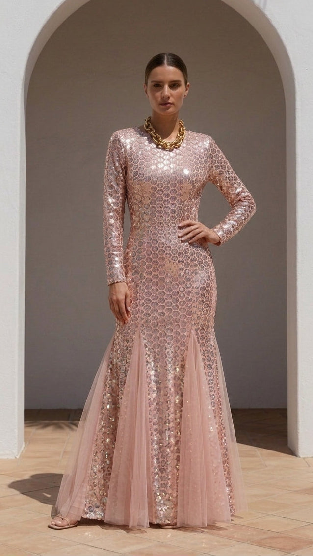 A woman models a long-sleeved, floor-length pink gown covered in shimmering sequins, standing in front of a white stucco archway with a tiled floor.