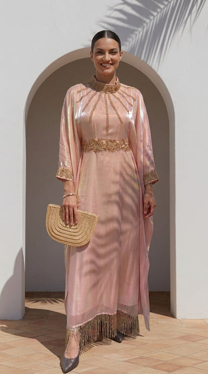 A woman models a light pink traditional long dress with gold embroidery, holding a woven straw clutch bag in front of a white arched doorway.