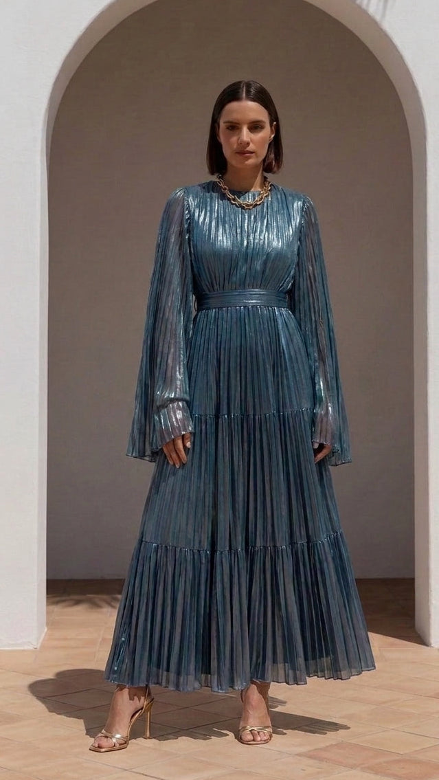 A woman stands in front of a white stucco archway wearing a long-sleeved, pleated blue maxi dress with a textured, shimmering finish. She is wearing clear high-heeled sandals and a chunky gold necklace.
