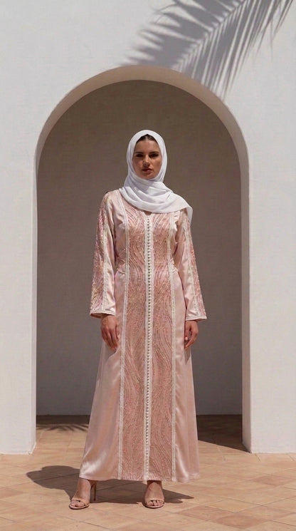 A woman wearing a light pink embroidered abaya and a white hijab stands in front of a white stucco archway with palm leaf shadows.