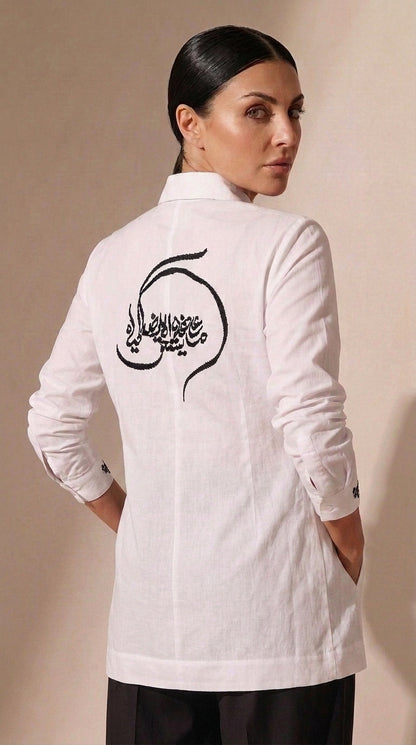 A woman shown from the back wearing a white long-sleeve tunic-style top with black Arabic calligraphy printed across the upper back, paired with dark pants, against a plain beige studio background.