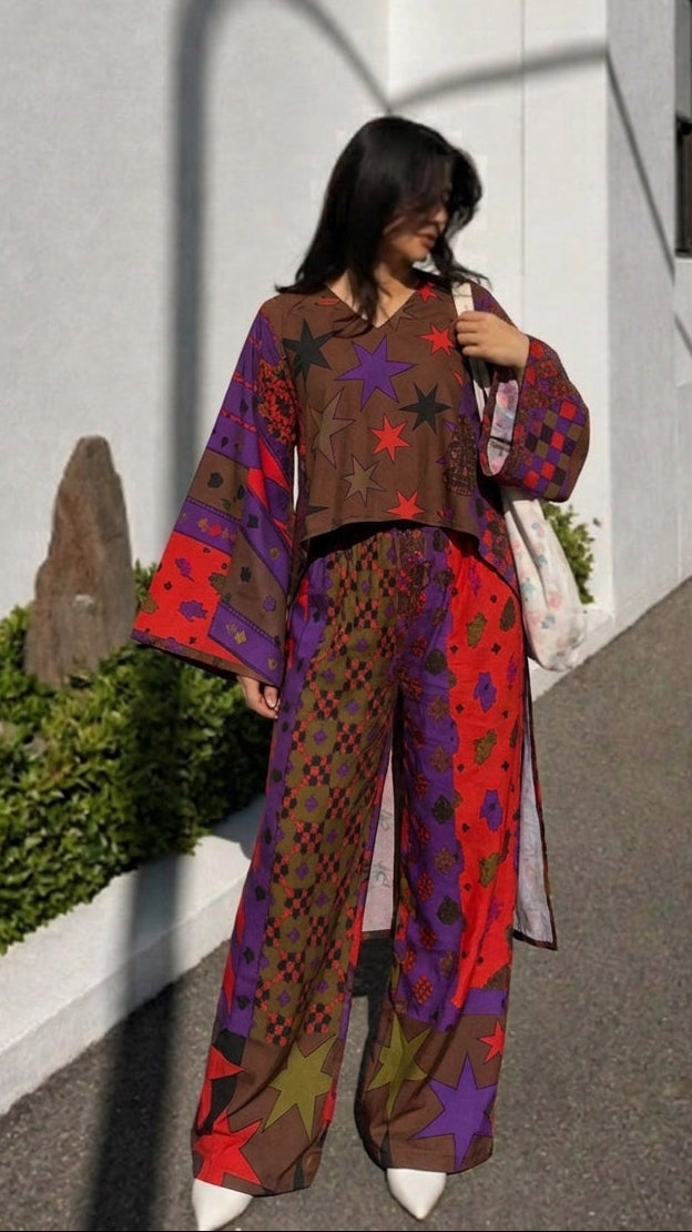 A woman stands outdoors on a paved walkway next to a white building, wearing a loose-fitting bohemian-style two-piece outfit with wide sleeves and wide-leg pants in brown, red, purple, and green with star and geometric patterns.