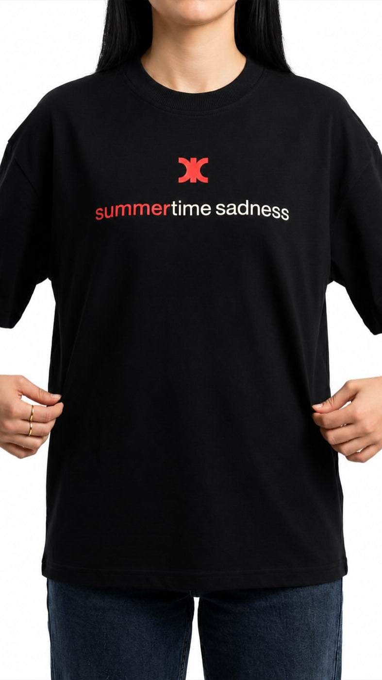 A person wearing a black short-sleeve T-shirt with a red and white 'summertime sadness' graphic on the chest, paired with dark blue jeans, against a plain white background.