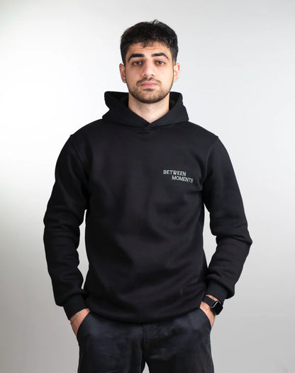 Person wearing a black hoodie with 'Between Moments' logo on a plain background