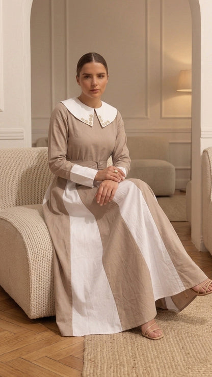 A woman sits on a cream upholstered armchair in a neutral-toned room, wearing a long beige dress with a white Peter Pan collar and white front panel.