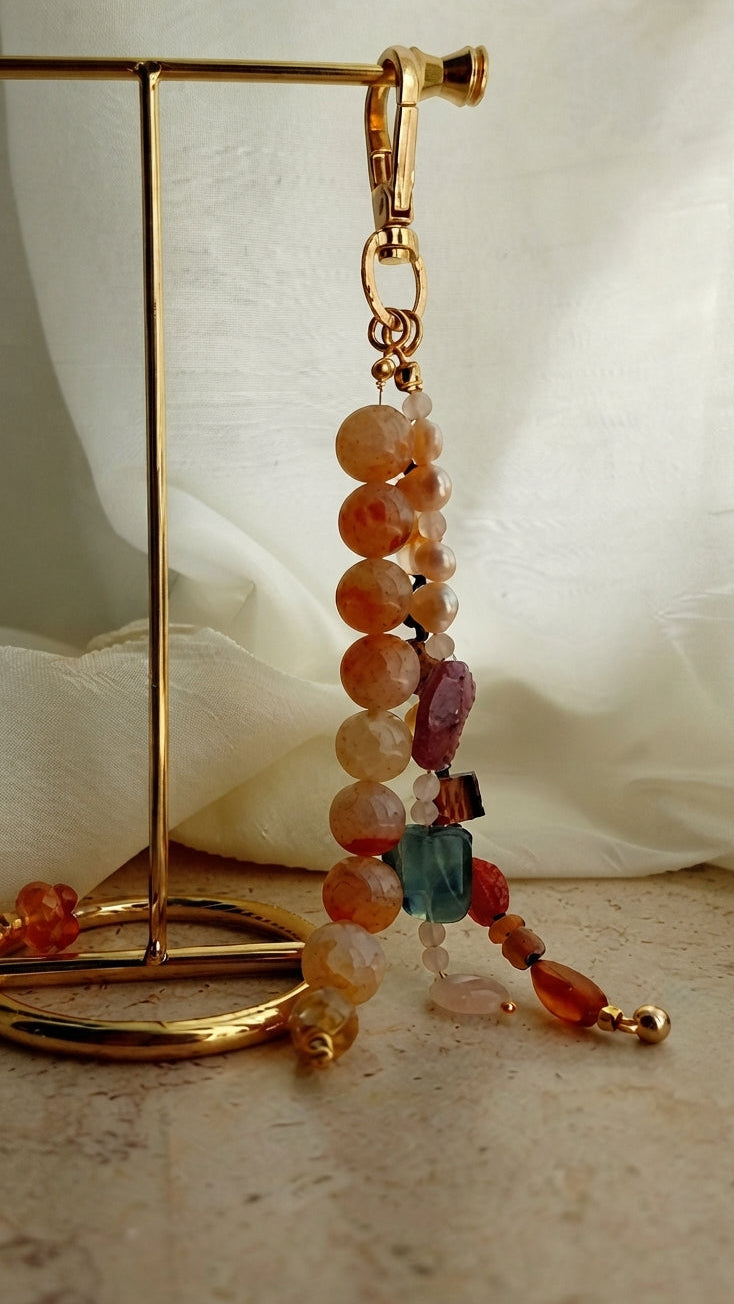 A beaded dangle earring with a gold-tone clasp, featuring round and faceted beads in orange, peach, pink, purple, and turquoise, displayed on a gold stand against a soft white fabric backdrop.