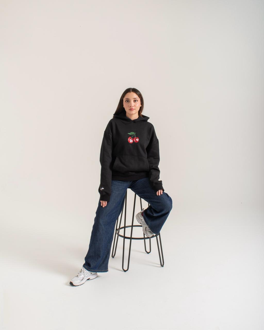 Person wearing a black hoodie with a cherry design, sitting on a stool against a white background