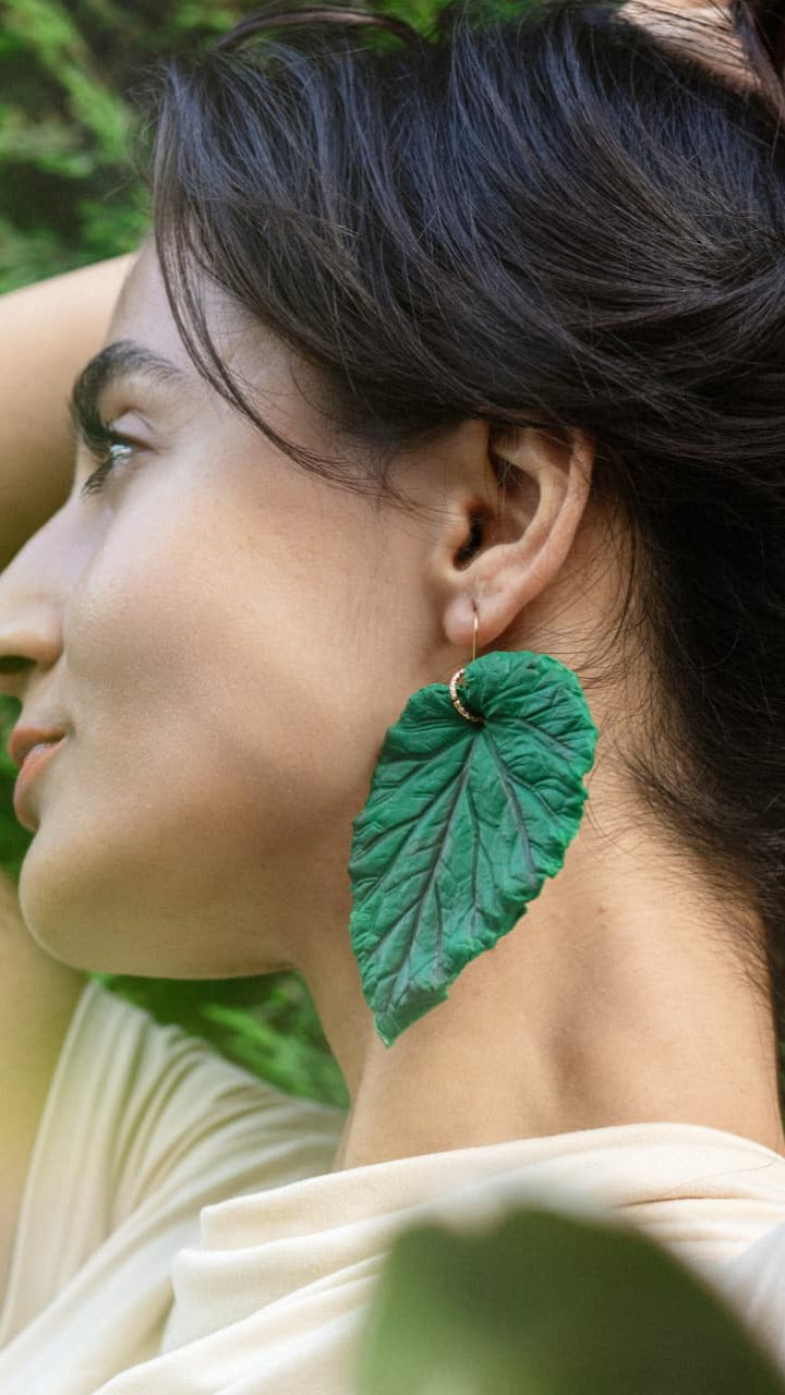 A close-up side profile of a woman wearing a large, textured green leaf-shaped dangle earring with a small gold hoop behind it, set against a soft-focus green outdoor background.
