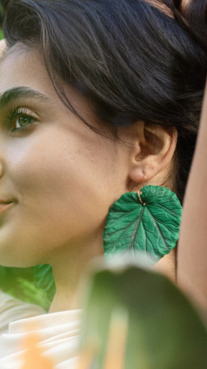 A close-up lifestyle shot of a woman wearing a large, textured green leaf-shaped dangle earring with a gold hoop.
