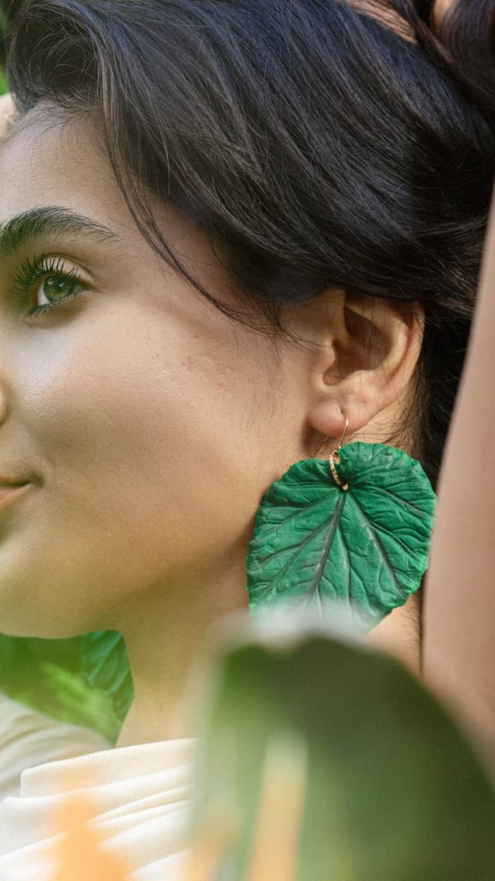 A close-up lifestyle shot of a woman wearing a large, textured green leaf-shaped dangle earring with a gold hoop.