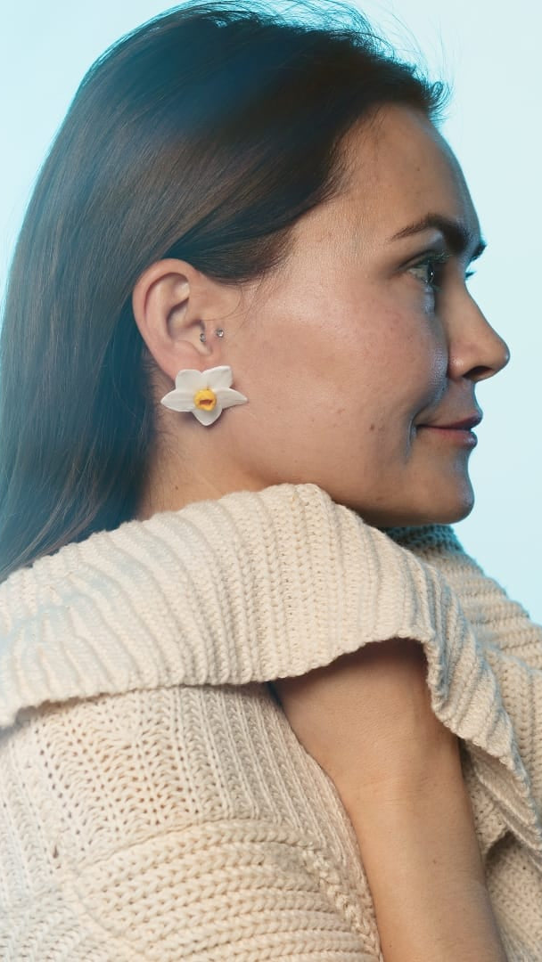 A woman in profile wearing a cream knit sweater and a white flower-shaped earring with a yellow center, set against a light blue background.