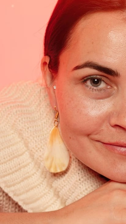 A close-up portrait of a woman with dark hair, wearing a cream knit sweater and large cream tassel earrings, resting her chin on her hands against a solid pink background.