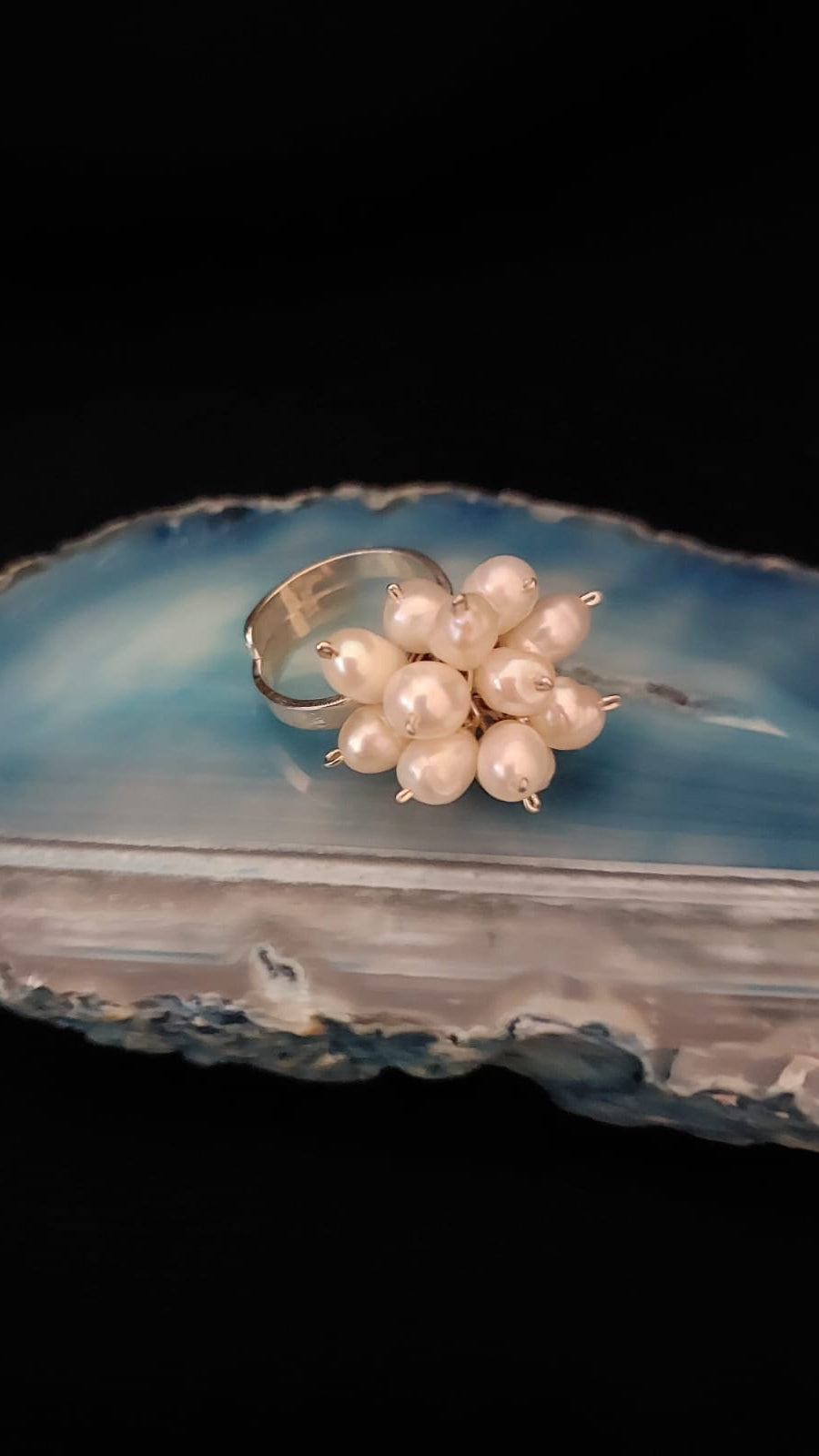 A silver ring featuring a cluster of small, lustrous white pearls, displayed on a polished blue agate slice against a solid black background.