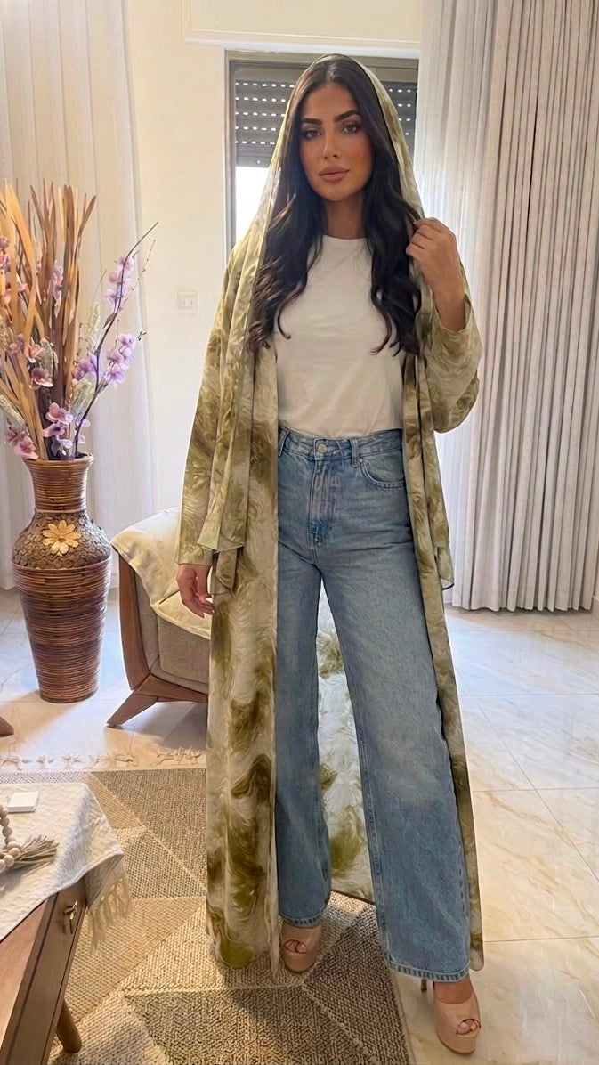 A woman stands indoors wearing a long, lightweight hooded cardigan with a beige, brown, and olive green tie-dye pattern over a white top and light blue wide-leg jeans.