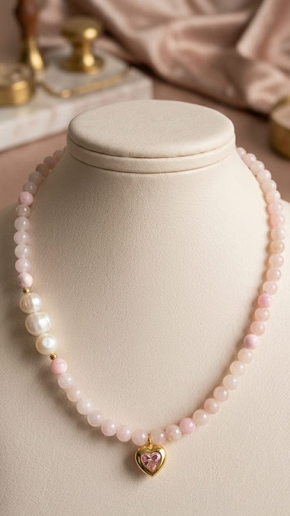 A delicate necklace displayed on a cream jewelry bust, featuring light pink and white beads with a small gold heart charm set with a pink stone.