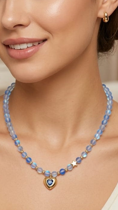 A woman wearing a delicate beaded necklace with light blue beads, a small heart-shaped charm with a blue center, and gold star accents, paired with a cream ribbed top.