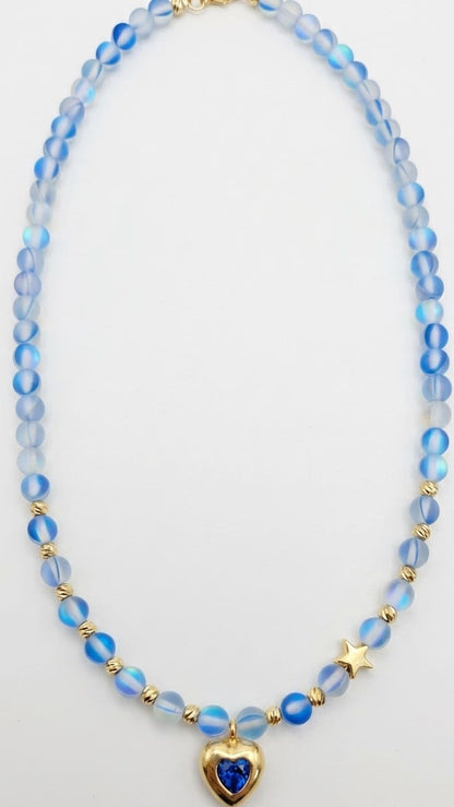 A beaded necklace featuring light blue and white marbled beads, accented with small gold spacer beads, a gold star charm, and a central gold heart pendant containing a deep blue heart-shaped stone, displayed on a clean white background.