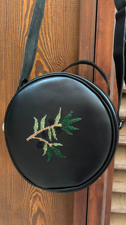 A round black faux-leather crossbody bag with an embroidered olive branch motif, hanging against a wooden door.