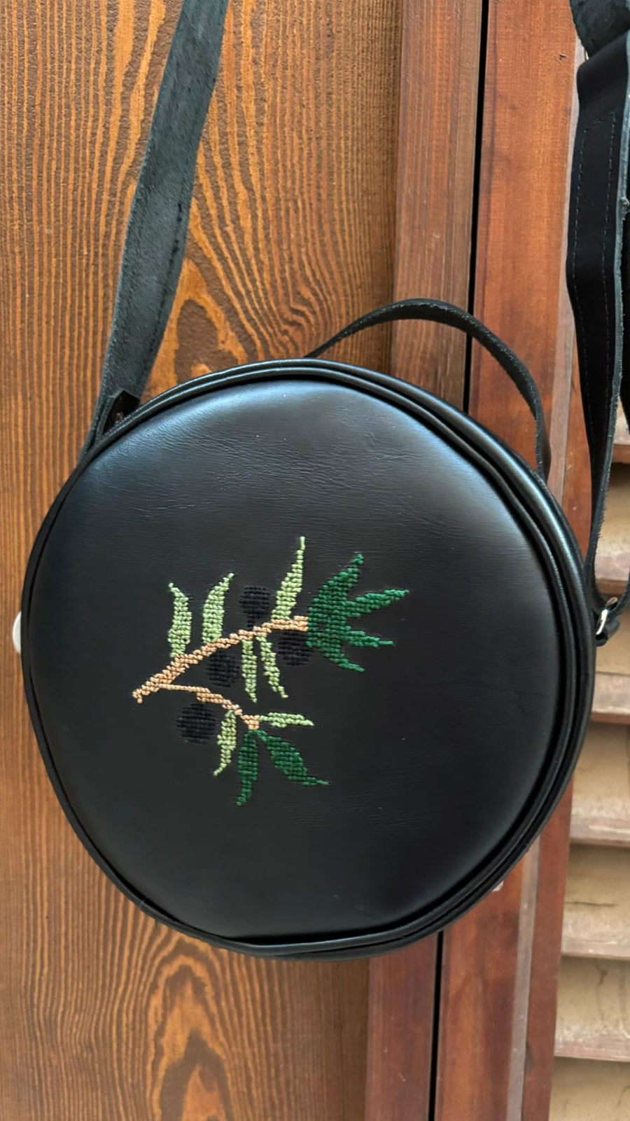 A round black faux-leather crossbody bag with an embroidered olive branch motif, hanging against a wooden door.