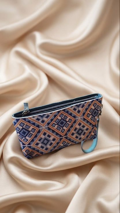 A small zip pouch with a geometric woven pattern in navy blue, orange, and beige, resting on draped cream satin fabric.