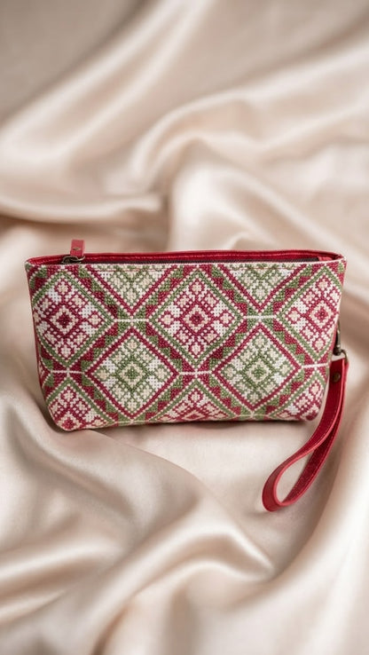A small zip pouch with a red wrist strap, featuring a geometric diamond pattern in red, green, and cream, styled on draped satin fabric.