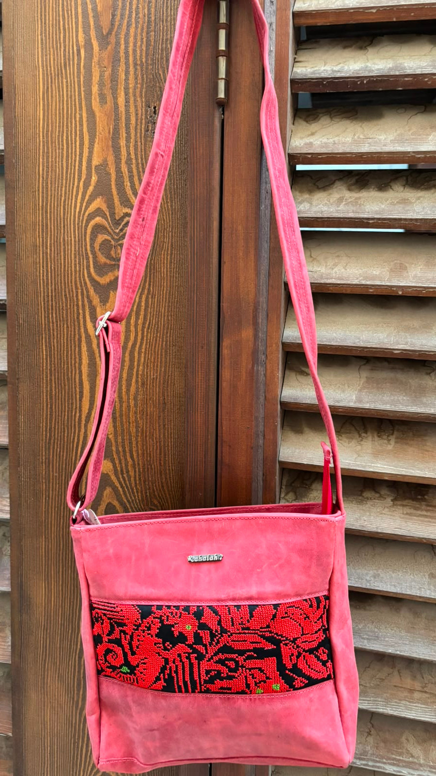 A pink crossbody bag with a red and black floral patterned front pocket, hanging against a wooden slat door.