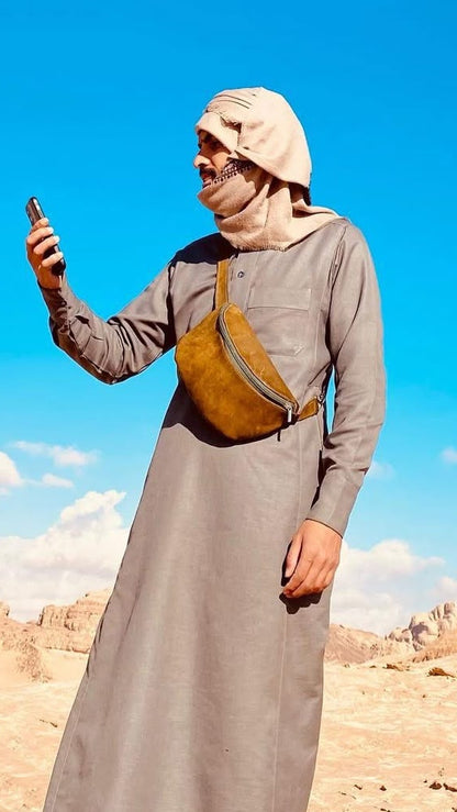 A man in a gray long-sleeve robe and beige headscarf stands in a desert landscape under a bright blue sky, holding a phone. He has a brown crossbody bag slung across his chest.