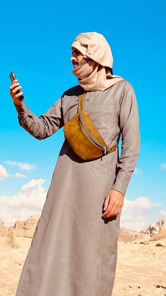 A man in a gray long-sleeve robe and beige headscarf stands in a desert landscape under a bright blue sky, holding a phone. He has a brown crossbody bag slung across his chest.