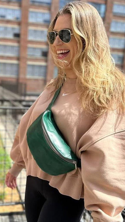 A woman wearing aviator sunglasses, a beige sweatshirt, and black leggings is pictured outdoors on a balcony, wearing a dark green crescent-shaped belt bag with a shiny mint green interior. A brick building is visible in the background.