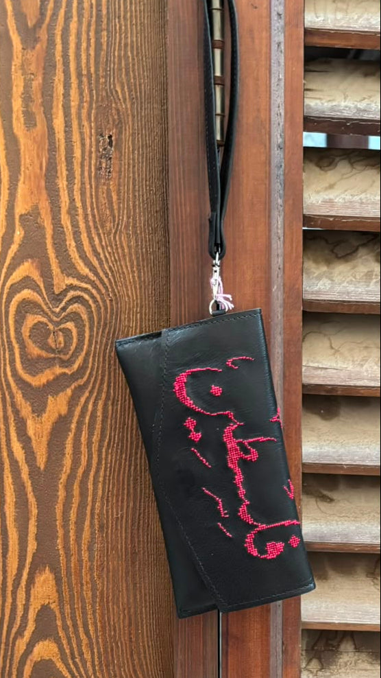 A black wristlet pouch with a pink gothic-style graphic hanging from a black wrist strap, set against a rustic wooden door background.