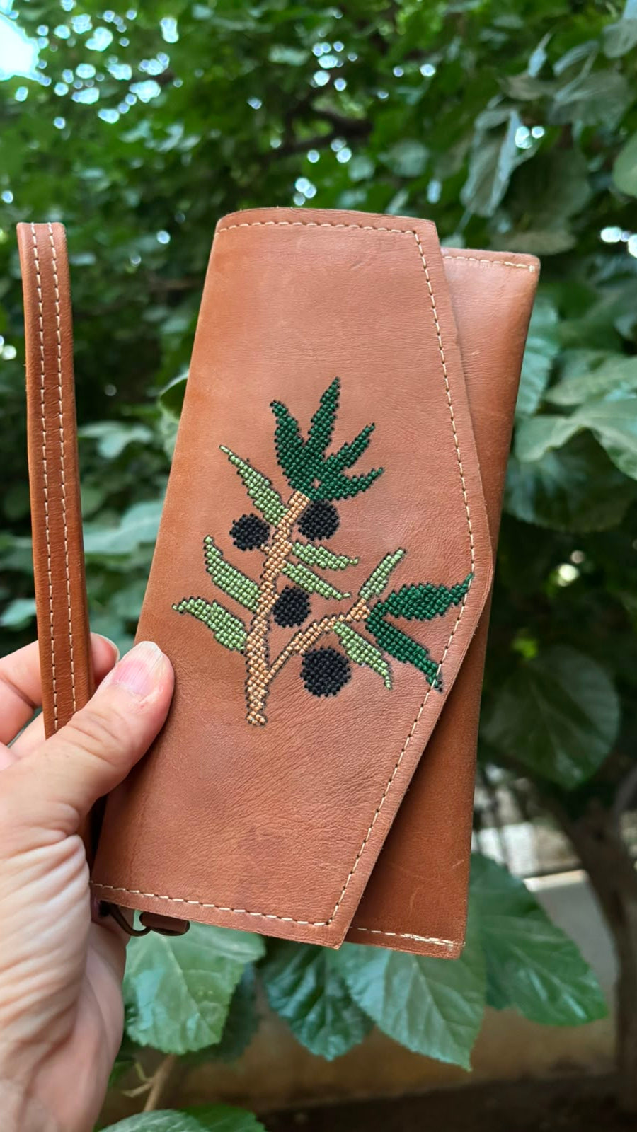 A tan leather wallet with an embroidered olive branch design, held by a hand against a background of green outdoor foliage.