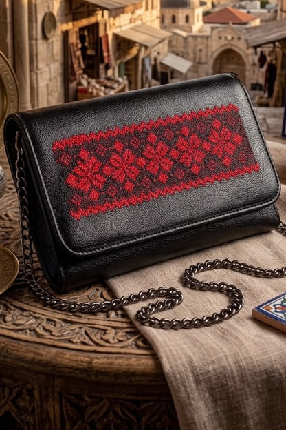 A black leather shoulder bag with a red floral embroidered pattern across the front flap, resting on a carved wooden table with a beige cloth and a metal chain strap. The background shows a scenic view of Jerusalem's old city with domes and arches.