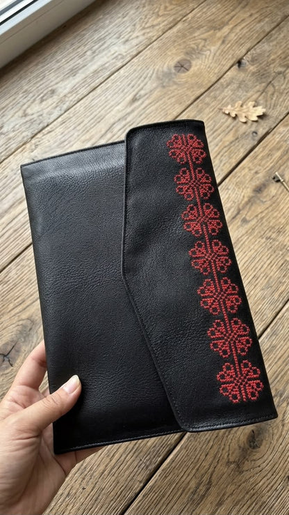 A black leather clutch or wallet with a vertical red embroidered pattern, held by a hand over a rustic wooden plank floor.