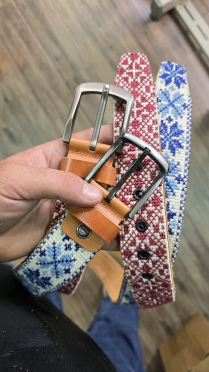 A hand holding three woven belts with silver buckles. The belts feature a tan leather base with blue and white geometric patterns on one, a red and white pattern on another, and a tan leather belt with a silver buckle on the third.