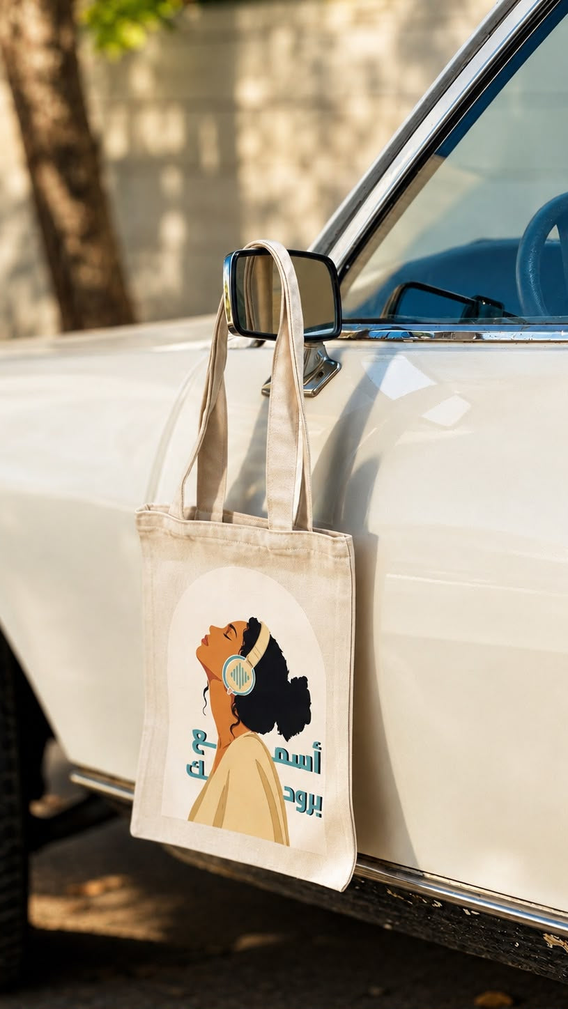 A canvas tote bag with a retro-style illustration of a woman wearing headphones and the text 'Bloom like a flower' hanging from the side mirror of a white vintage car.