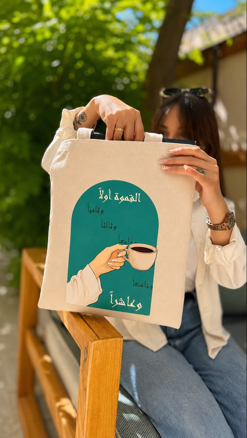 A woman sitting on a wooden bench outdoors holds up a cream canvas tote bag with a teal graphic print featuring Arabic text and an illustration of a hand holding a coffee cup.
