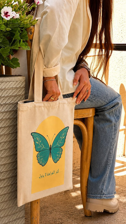 A person sitting on a wooden stool holds a cream canvas tote bag with a teal butterfly and yellow sun graphic, next to a potted plant.
