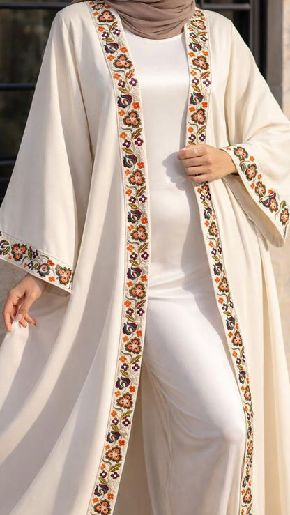 A woman wearing a long cream abaya with colorful embroidered borders, a matching white inner dress, and a taupe hijab, standing in front of a beige wall and window.