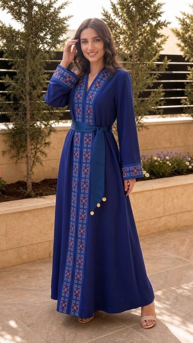 A woman wearing a long royal blue caftan dress with intricate red and white geometric embroidery, standing in an outdoor patio setting with plants and stone walls.