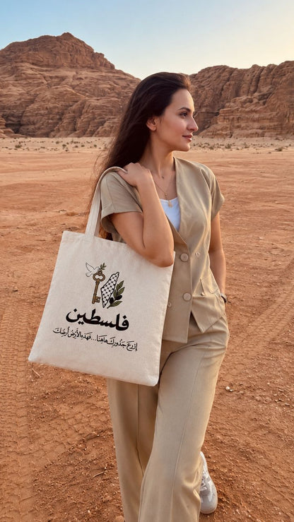 A woman in a beige suit walks across a desert landscape carrying a cream canvas tote bag printed with a bee logo and Arabic text. The background features reddish-brown rock formations under a clear blue sky.