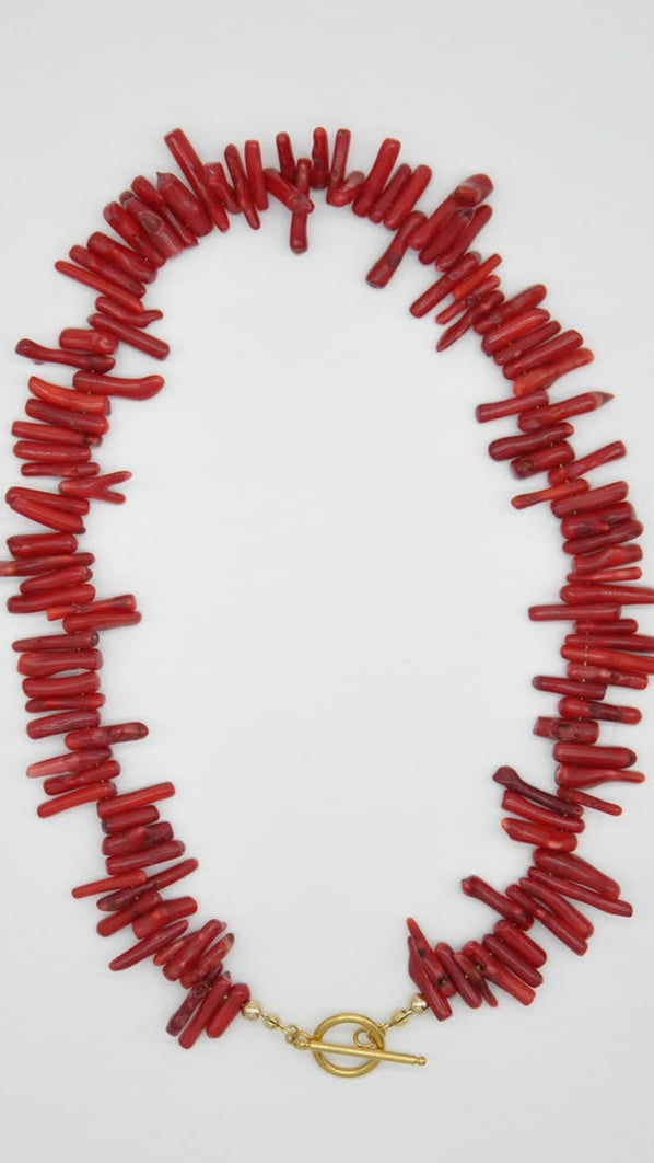 A chunky red coral-style necklace with irregular cylindrical beads and a gold toggle clasp, laid flat on a light gray background.