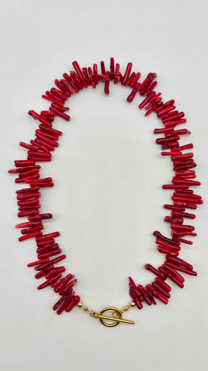 A red coral-style chunky necklace with a gold toggle clasp, laid flat on a light gray background.